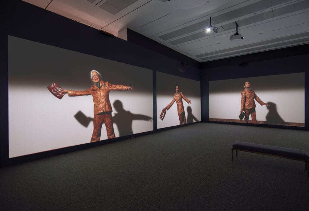 Two parallels walls with projected images of three women wearing bejewelled suits, holding clutch persons. They all have their arms outstretched.