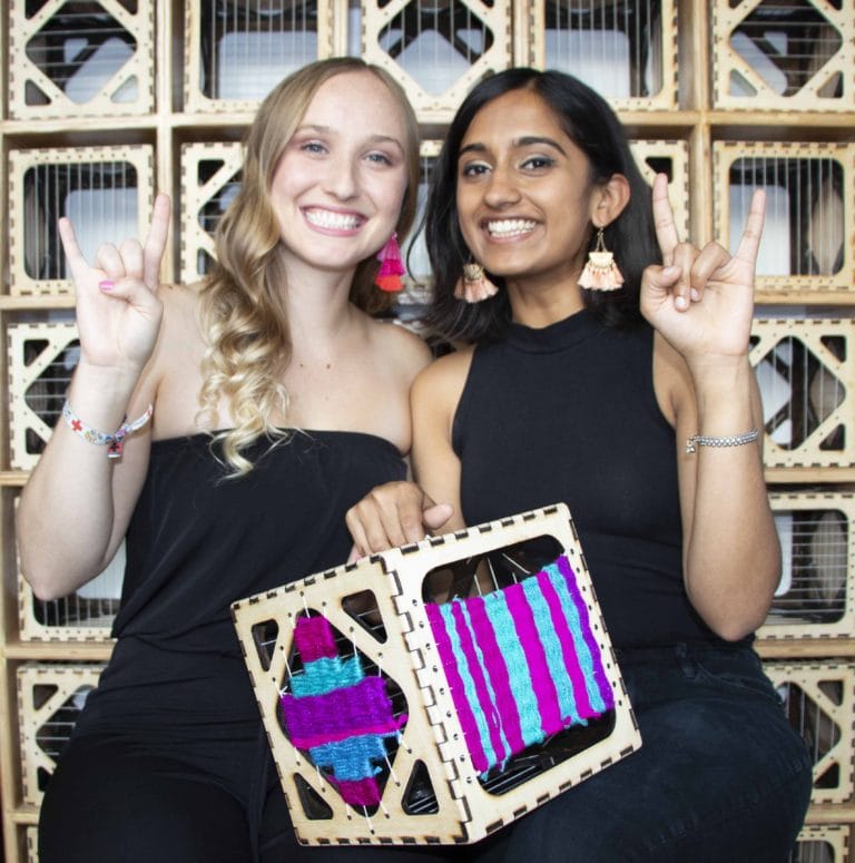 two young woman with a weaving project between them both smiling and with the hookem sign with their hands
