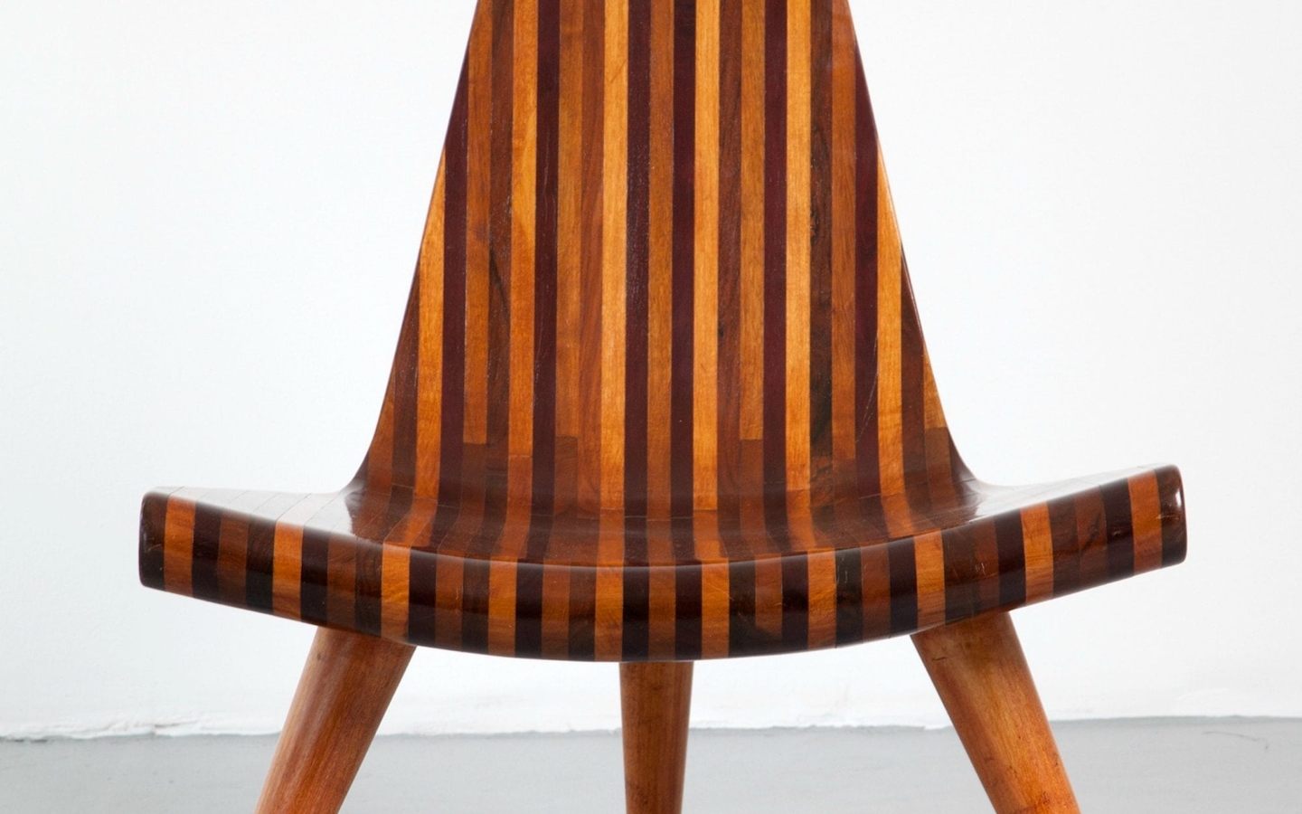 A wooden chair with three legs. The wood within the chair is arranged in thin vertical stripes of varying shades of brown. The seat of the chair is wide and transitions seamlessly into the back of the chair, which tapes to a peak.