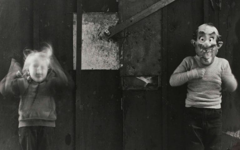 Black and white image of two people facing the viewer, against a wooden wall. On the left, a child jumps up, arms blurred by motion. On the right, a figure wears a mask depicting a smiling balding man with a mustache, wide eyes, and crooked teeth.