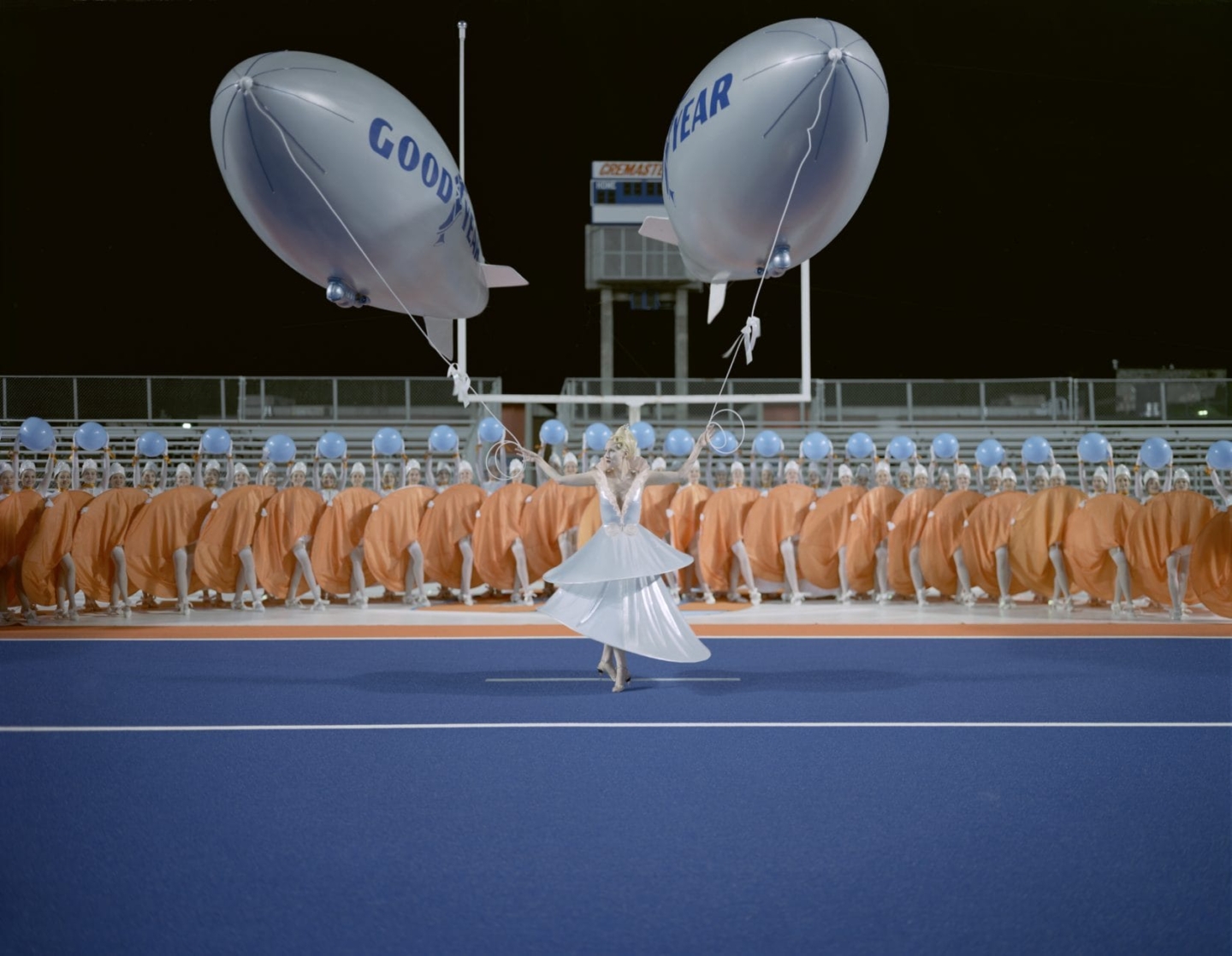 In the foreground, a blonde woman wearing a futuristic pale blue shiny dress stands on blue AstroTurf, holding a large GoodYear blimp balloon in each hand. Behind her stands a line of women in similar dresses, colored orange, holding up blue balls in their arms. Behind them are metal bleachers. It is nighttime.