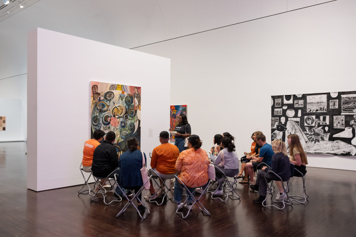 A group of people sit on foldable stools in an art gallery, listening to a speaker talk about a painting on the wall.