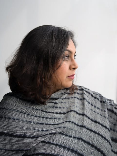 A person with shoulder-length dark hair, wearing a grey and black striped shawl.