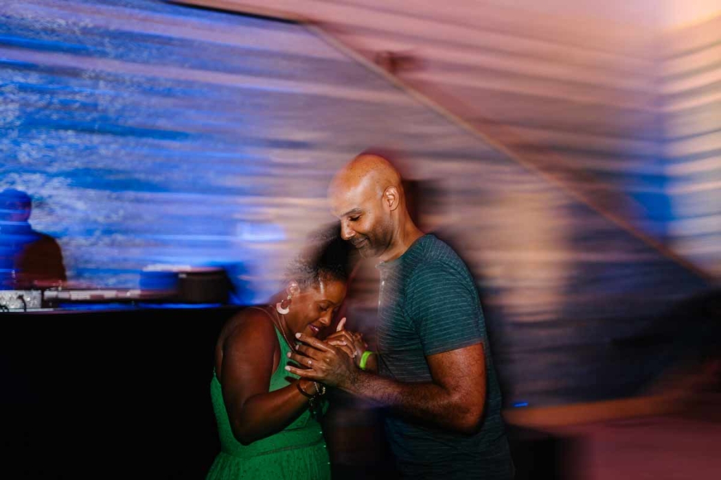 Two people dancing closely together in a lively setting with blue and purple lighting. The background includes a DJ booth and a person standing behind it, capturing a moment of intimacy and connection.
