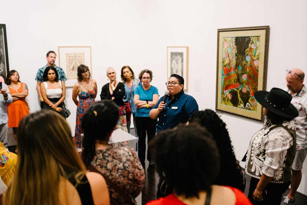 A group of people gathered in an art gallery or museum, listening to a person who is speaking into a microphone. The speaker is wearing a dark blue shirt and a name tag. The attendees are standing around, some with their arms crossed, looking at the speaker or the artworks on the walls. Several framed artworks are visible, including a colorful painting and a monochrome piece. The setting suggests an art tour or gallery talk.
