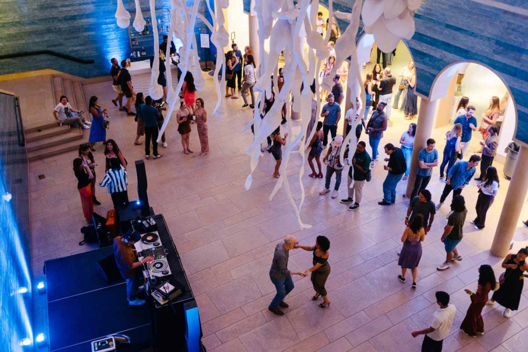 A lively indoor event where people are socializing, dancing, and enjoying music. The hall has a high ceiling, decorated with white streamers. A DJ booth is in the lower left corner, and colorful blue and yellow lighting creates a festive atmosphere. The architecture includes arches and a staircase.