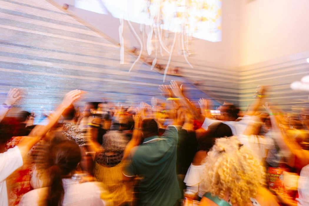 A large group of people with their hands raised, gathered indoors, possibly at a concert or festival. The scene is dynamic with a sense of movement and energy. The background features a blue and white striped wall and hanging decorations.
