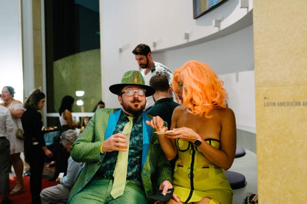 A group of people at a social event or gathering. The focus is on two individuals in the foreground, one in a green suit with a matching hat, holding a drink, and the other with bright orange hair in a yellow dress, holding a small plate of food. The background includes other attendees, some standing and some sitting, engaged in conversation.