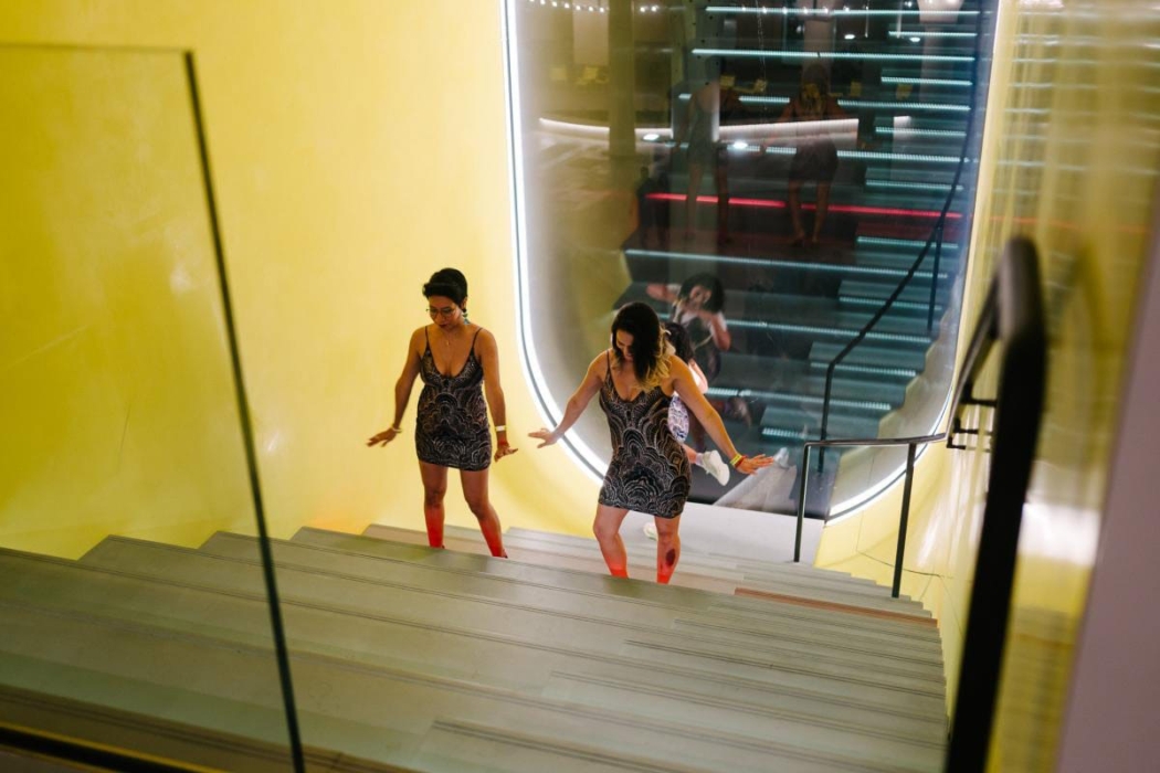 Two individuals walking up a set of stairs, enclosed by a yellow wall on one side and a glass barrier on the other. They are wearing matching dresses with a black and white pattern and red stockings. The background features a modern, well-lit interior with another set of stairs visible through the glass barrier.