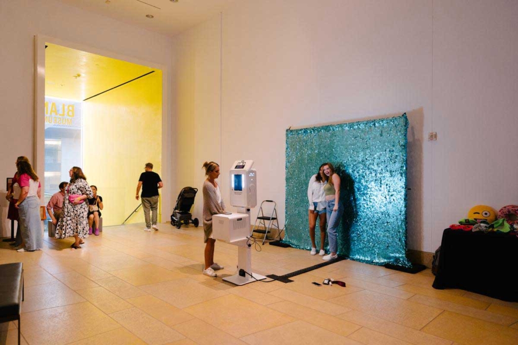 Several people are inside a museum or public space, some standing and some sitting. On the right side, there's a photo booth with a blue sequin backdrop, with two people posing in front of it. A table with props like an emoji pillow is visible. In the background, a yellow-lit entrance with 'BLANK' partially visible suggests the name of the exhibit or museum. A stroller is near the entrance, and the floor is tiled.