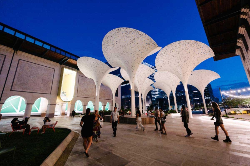 A modern outdoor area with large, white, flower-like structures illuminated from below. These structures are supported by tall columns, creating a canopy over the public space. The background features a building with geometric patterns and lit arched openings, indicating a vibrant evening or night scene. People are walking or sitting in the area, adding to the lively atmosphere