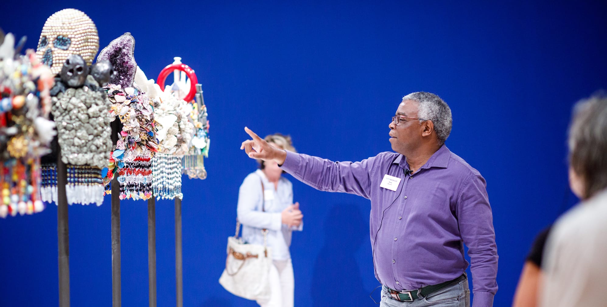 A male gallery teacher gestures to colorful, sculpture like artworks on poles.
