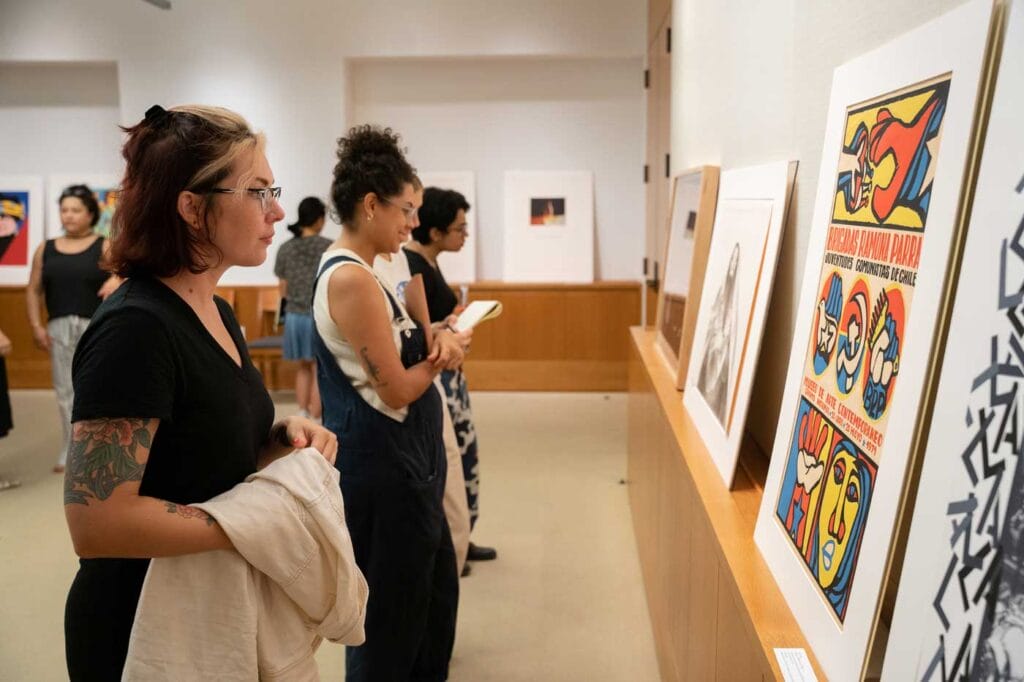 A group of people in an art gallery, observing various pieces of artwork displayed on the walls and on a shelf. The artworks include colorful illustrations and black-and-white photographs. One prominent piece on the right side is a colorful poster with text that reads 'BRIGADAS RAMONA PARRA JUVENTUDES COMUNISTAS DE CHILE' and features stylized faces and hands in red, blue, and yellow colors. The setting appears to be a well-lit, modern gallery space with wooden accents. The people are engaged in viewing and possibly discussing the art, indicating an interest in the exhibits.