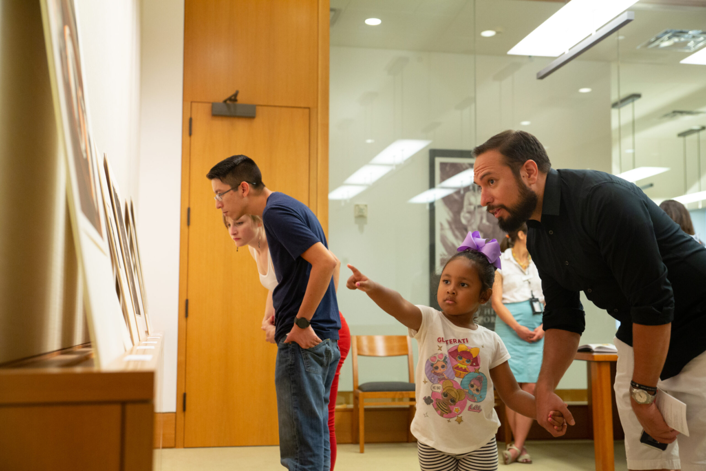 A group of people in a gallery. A young girl is pointing at something on the wall, with an adult bending down next to her. Another person closely examines the items on display. The room has wooden furniture and a glass wall in the background, capturing a moment of engagement and curiosity.