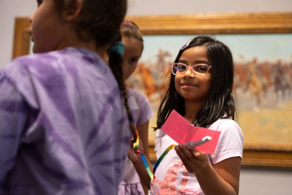 A group of children in a room with a framed painting in the background. One child in the foreground holds a pink piece of paper and a small foil-wrapped object. The children wear casual clothing, and the child in the foreground has glasses and long hair. The painting in the background depicts a scene with horses and riders. 