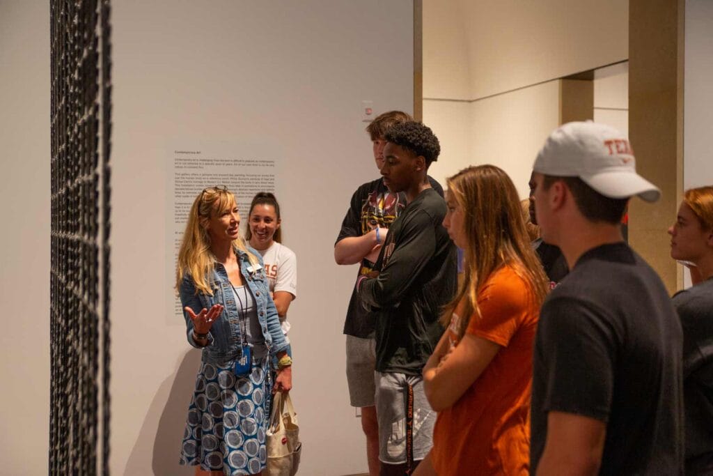 "A group of people standing in an art gallery or museum, listening to a woman who appears to be a guide or instructor. They are gathered around an art piece, with a text panel on the wall behind them that contains information about the exhibit. The text on the wall discusses contemporary art and its various themes and approaches.