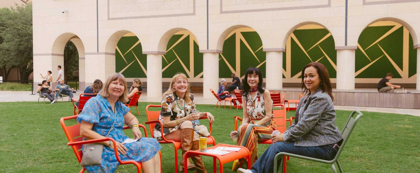 A group of people sitting on bright orange and green chairs arranged in a circle on a grassy area. The outdoor courtyard features a building with arched openings and green geometric designs on the walls in the background. Additional people are seen in the background, some sitting and some standing, engaging in various activities. This scene suggests a casual, social gathering or event.