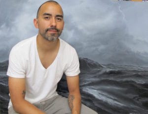 A person sitting in front of a backdrop depicting a stormy sea with dark, turbulent waves and a cloudy sky. The individual wears a white t-shirt and gray pants.