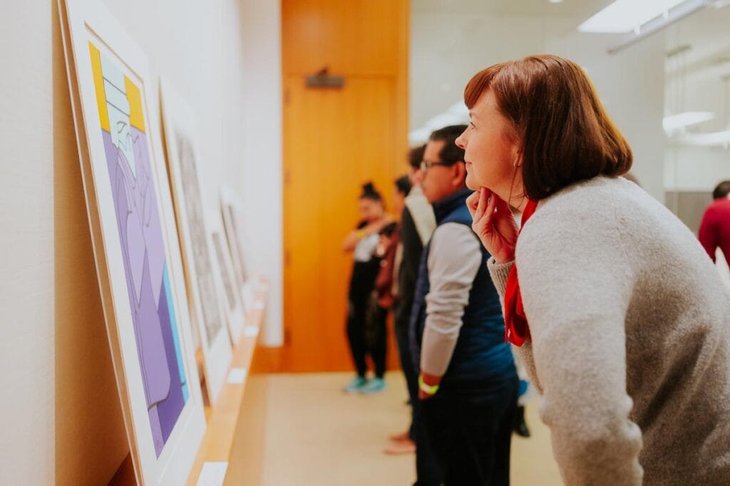 A group of people in an art gallery, engaging with framed artworks displayed on a shelf along the wall. One person closely examines a piece, with others casually observing. The gallery setting is well-lit and showcases a mix of abstract and monochromatic art styles, highlighting the cultural and educational value of art exhibitions.