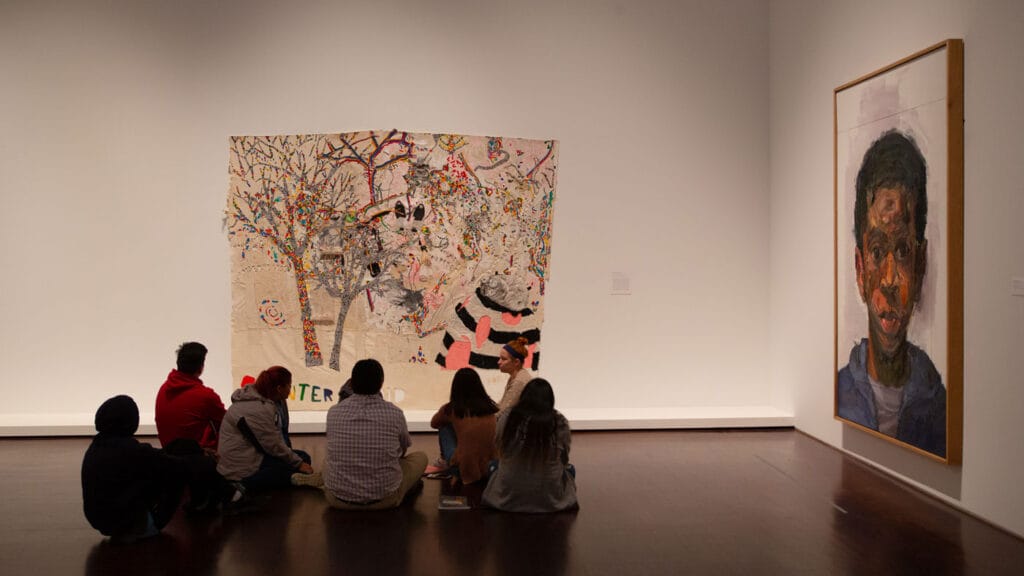 A group of kids is sitting on the floor in an art gallery, observing two large artworks displayed on the walls. The artwork on the left is a colorful, abstract piece featuring trees and various shapes and patterns. The artwork on the right is a large portrait of a person with a serious expression, painted in a realistic style with visible brushstrokes. The gallery has a clean, minimalist design with white walls and a polished wooden floor. The scene captures a moment of art appreciation and education.