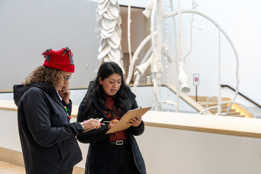Two individuals are standing indoors, one holding a clipboard and pen, while the other points at the clipboard. They appear to be engaged in a discussion or review. The background features a modern, artistic installation with white, abstract structures and a staircase.