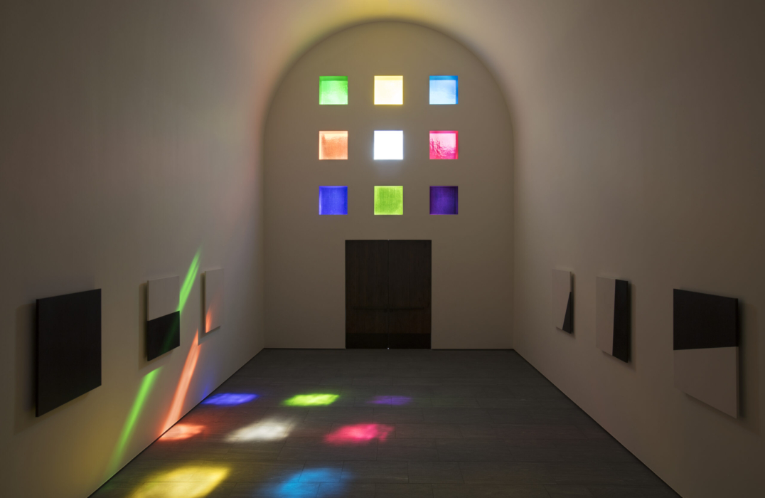 Interior view of Ellsworth Kelly's Austin; the image faces the entrance at a time of day when the sun shining through the nine stained glass blocks create rainbow shadows on the wall and floor.