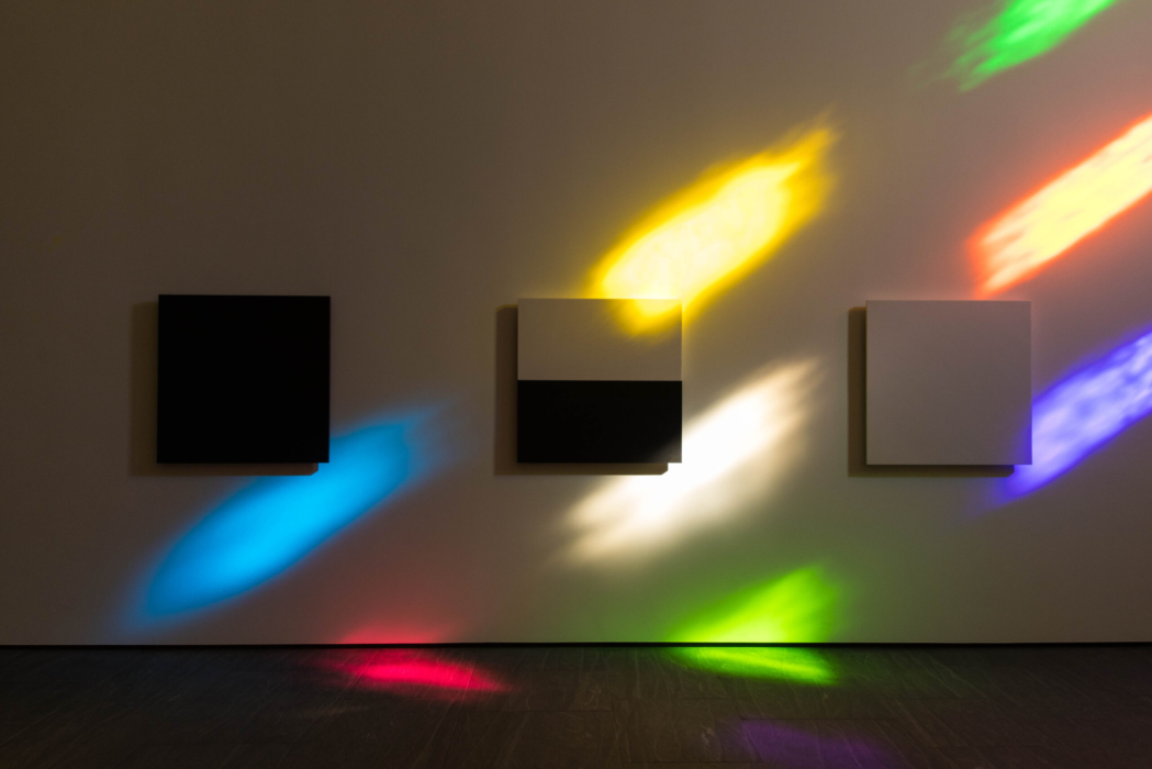 Interior wall shot of Austin by Ellsworth Kelly. Three marble panels line the wall as square rainbow colored shadows drape the wall. The panel on the far left is solid black, the panel in the middle is half white on top and half black on the bottom, and the far right panel is solid white.