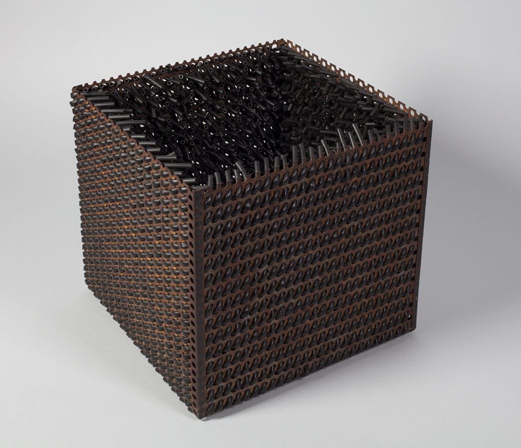 A sculpture of a metal cube with the top open. The outside appears with a woven texture, and the inside contains small metal tubes reaching in from the sides at varous angles.