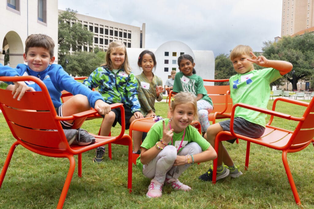 A group of smiling young students sit in chairs on the Blanton grounds. Austin by Ellsworth Kelly is in the background. 