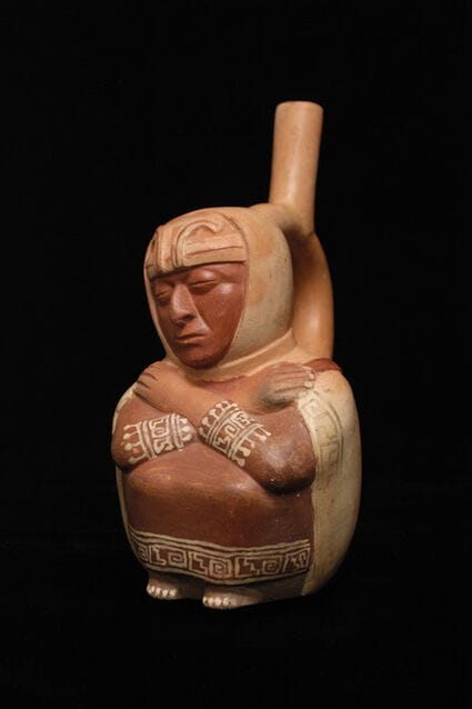 Stirrrup spout of blind figure. The ceramic figure is painted in shades of brown and is crouched down with their arms crossed over their chest.