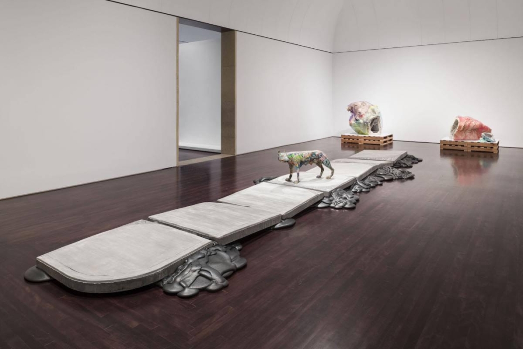 The image shows an art installation in a gallery space. The installation features a pathway made of large, flat, rectangular slabs with a colorful animal sculpture, possibly a dog or a cat, positioned on top of the slabs. Surrounding the slabs are irregular, organic shapes that appear to be made of a different material, possibly metal or resin. In the background, there are two large, abstract sculptures placed on wooden pallets. The gallery has white walls and a dark wooden floor, creating a stark contrast with the art pieces. This image is interesting because it showcases a contemporary art installation that combines different materials and forms, creating a visually engaging and thought-provoking environment.