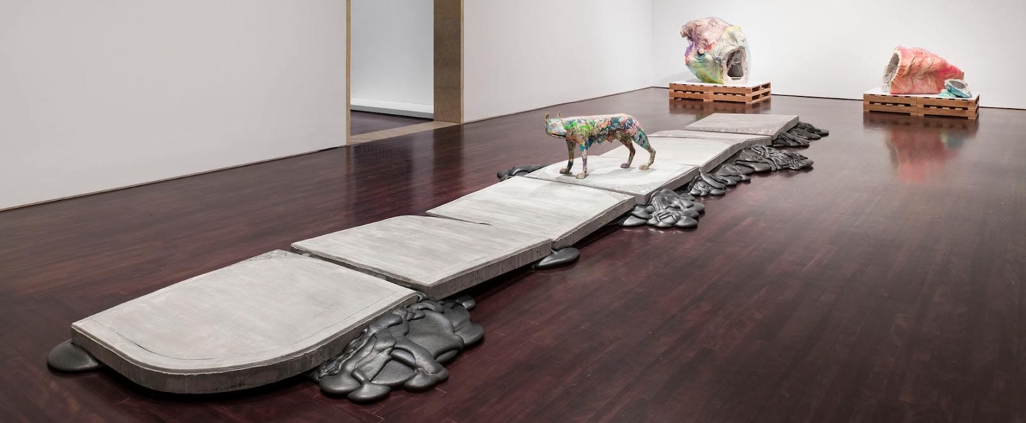 An art installation in a gallery space featuring a series of large, flat, rectangular slabs arranged in a line on the floor. On one of the slabs stands a colorful, abstract sculpture of an animal, possibly a dog or a wolf. The slabs are surrounded by dark, amorphous shapes that resemble melted material. In the background, there are two large, colorful sculptures that appear to be abstract representations of heads or skulls, each placed on wooden pallets. The gallery has a polished wooden floor and white walls, creating a clean and modern setting for the artwork