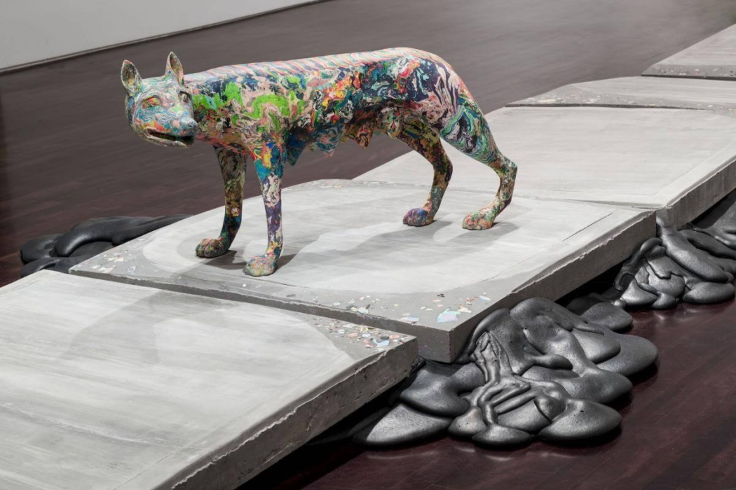 A colorful, abstract sculpture of an animal, possibly a wolf or a dog, standing on a series of large, flat, rectangular concrete slabs. The sculpture is covered in vibrant, swirling colors. The concrete slabs are arranged in a path-like manner, with dark, organic shapes appearing to ooze or melt from underneath the slabs. The setting is an art gallery with a polished wooden floor, creating a contrast between the vibrant sculpture and the industrial concrete slabs.