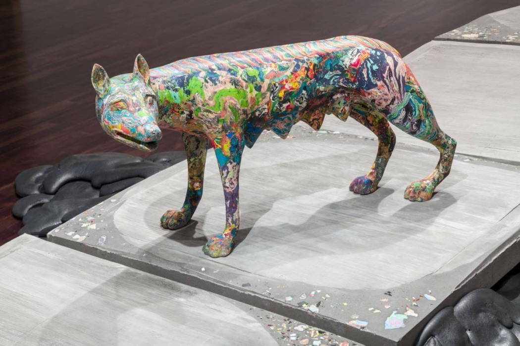 A colorful sculpture of an animal, likely a wolf or dog, standing on a platform. The sculpture is covered in a vibrant, multicolored pattern resembling paint splatters and swirls. The platform beneath it is gray with scattered paint splatters around it. The background features a dark wooden floor and some black, cloud-like shapes on the left side of the platform. The unique and vivid design of the sculpture makes it an interesting piece of art.