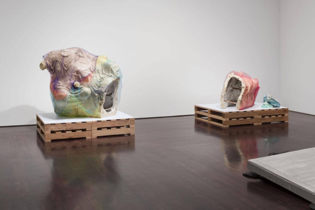 An art gallery or exhibition space with two large, abstract sculptures displayed on wooden pallets. The sculptures appear to be made of plaster or concrete with colorful, translucent overlays. The larger sculpture on the left features a mix of pastel colors, while the smaller sculpture on the right has a red overlay and an additional smaller piece beside it. The gallery floor is polished dark wood, and the walls are plain white, providing a neutral background that highlights the sculptures. There is also a partial view of another platform in the bottom right corner of the image.