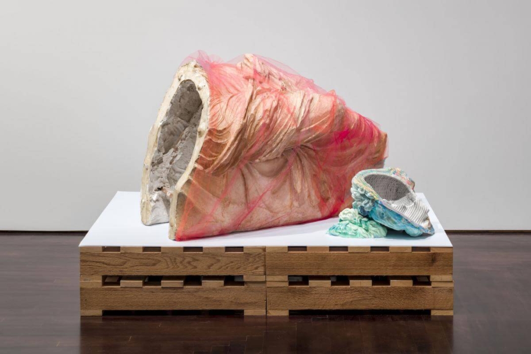 The image shows a sculptural artwork displayed on a wooden platform. The sculpture consists of two main parts: a larger, hollow, shell-like structure wrapped in a translucent pink material, and a smaller, similarly shaped object with a colorful, textured surface. The larger piece appears to be made of a rough, stone-like material, while the smaller piece has a more intricate, layered appearance. The artwork is set against a plain background, emphasizing the textures and forms of the sculptures. This image is interesting because it showcases a unique and abstract piece of art that combines different materials and textures, inviting viewers to interpret its meaning and significance.