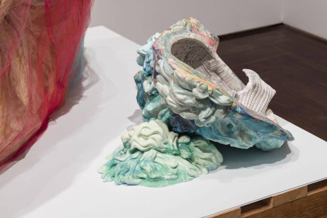 The image shows a colorful, abstract sculpture placed on a white surface. The sculpture appears to be made of various materials, including a textured, woven-like material and a smooth, painted surface. The sculpture has a dynamic, flowing form with a mix of pastel colors, including shades of blue, green, pink, and purple. The structure resembles a combination of organic shapes and possibly a shell or a piece of coral, with intricate details and a sense of movement. The left side of the image shows another part of the sculpture or a different piece with a red and beige fabric-like material. The artwork is displayed in a gallery setting with a wooden floor and white walls.