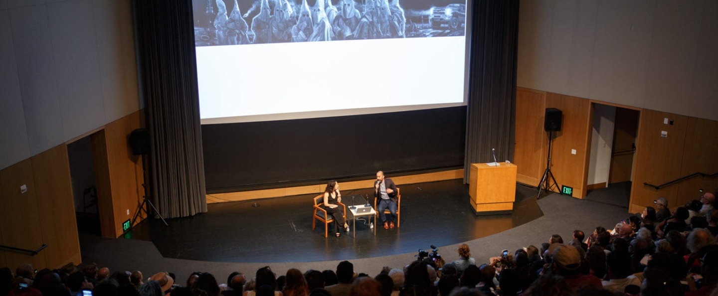 A lecture or discussion event in an auditorium with two speakers on stage, seated in front of a large screen displaying an image of individuals in white robes and hoods. The audience is attentively listening. The setting includes a podium, speakers, and exit signs.