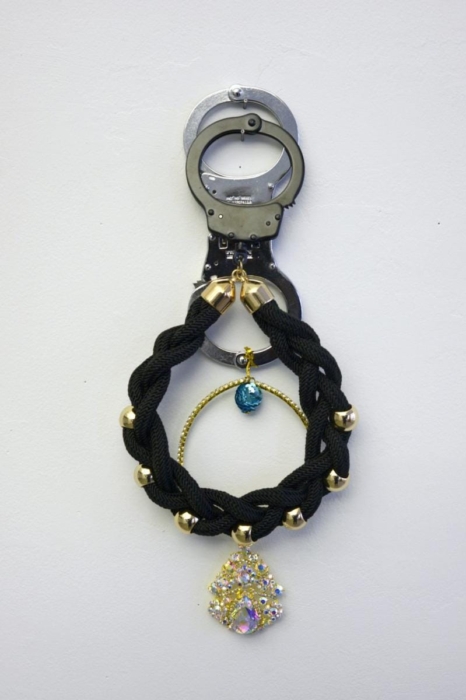 The image shows a pair of handcuffs with decorative elements attached to them. The handcuffs are metallic and standard in design. Attached to the handcuffs is a black braided cord with gold-colored beads interspersed along its length. Hanging from the braided cord is a gold-colored circular frame with a blue gemstone in the center. Below the circular frame, there is a large, ornate pendant adorned with multiple small, colorful gemstones. The combination of the utilitarian handcuffs with the decorative elements creates an interesting juxtaposition, blending themes of security and adornment.