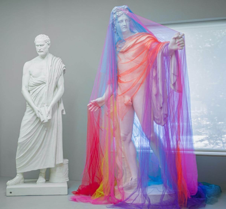 Two classical-style statues are displayed in an art gallery. The statue on the left is made of white marble and draped in a toga, holding a scroll or book. The statue on the right is also a white marble figure but is partially covered with a colorful, translucent fabric that drapes over the head and body, creating a striking contrast with the traditional marble. This modern fabric element adds a vibrant touch to the classical statue. The setting of the gallery emphasizes the blend of classical and contemporary art styles.