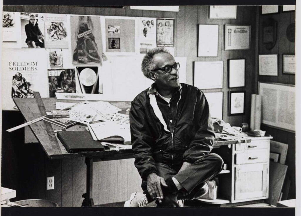 A black and white photograph of Charles White sitting on a desk in a cluttered workspace. He is wearing a jacket and glasses, with his legs crossed and hands resting on his knee. The desk is covered with various papers, books, and drawing tools. The background features a wall adorned with numerous posters, certificates, and framed pictures, including posters with the titles 'FREEDOM SOLDIERS,' 'EBONY,' and 'BLACK CAUCUS.' The workspace appears to be an artist’s or designer’s studio, with a focus on African American culture and history.