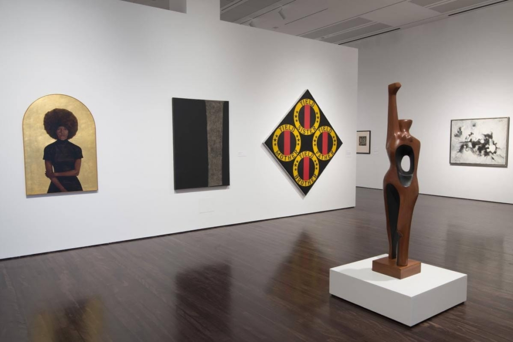 An art gallery featuring various artworks and a sculpture. On the left is a painting of a person with an afro, viewed from behind against a gold background. Next, a rectangular abstract painting with a black and gray stripe. Beside it, a diamond-shaped piece with 'MOTHER,' 'FATHER,' 'SISTER,' and 'BROTHER' in a circular pattern on a yellow background. A tall, hollow wooden sculpture stands on a pedestal in the foreground. On the far right, a black and white abstract painting. The gallery's modern, clean design with white walls and dark wooden floors highlights the diverse display.