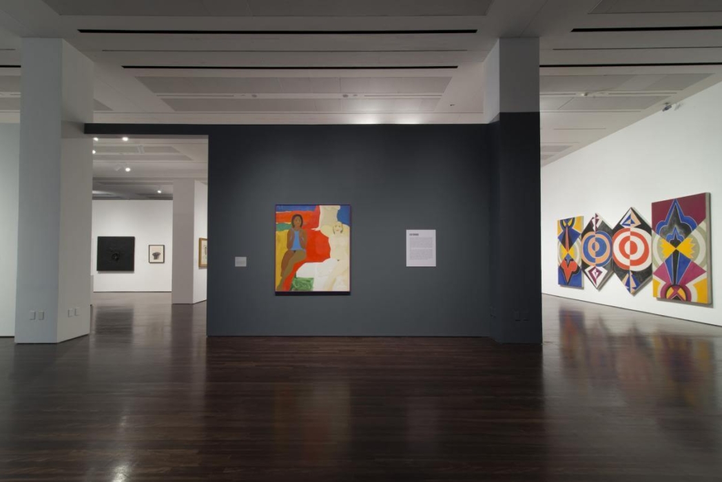 An art gallery with several paintings displayed on white walls and dark wooden floors. The central piece is a colorful painting of abstract human figures on a dark gray wall. To the right is a text panel about the artwork. Three large, colorful geometric paintings are arranged in a row on a white wall to the right. Additional artworks, including a black square painting and a framed drawing, are visible in the background.