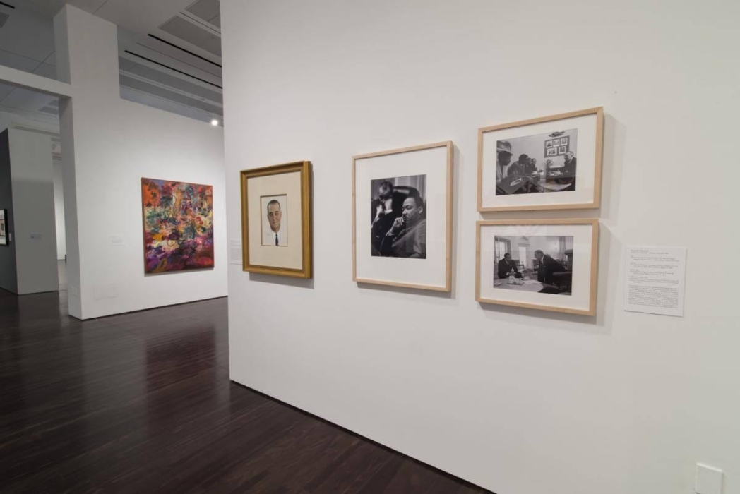 An art gallery with several framed artworks displayed on white walls. The right wall has four pieces: one vibrant, abstract color painting and three black-and-white photographs depicting various scenes, including people in different settings. A text plaque provides information about the artworks or artist.