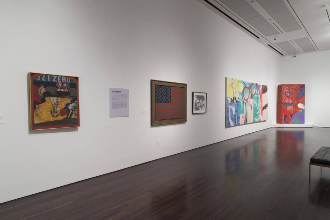 An art gallery with white walls and dark wooden floors, showcasing various artworks. The leftmost piece has the text '321-ZERO' at the top, featuring an abstract scene with vibrant colors. To the right is a plaque with information. Next, a framed piece resembles an American flag with blue and red stripes, followed by a black and white artwork. Further right, there's a large, colorful abstract painting. In the far corner is another vibrant painting with red and blue tones. A bench sits on the right for visitors.