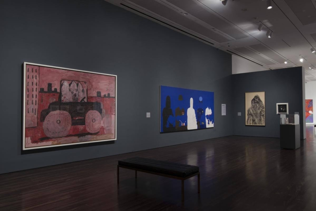 An art gallery with several paintings displayed on dark-colored walls and a wooden floor. A bench sits in the center for visitors. The paintings include a red and black vehicle with a cityscape background, a blue piece with silhouetted figures and a central white figure, a figure wrapped in a cloak or blanket, and a smaller framed piece on a pedestal. The gallery is well-lit with spotlights highlighting each artwork.