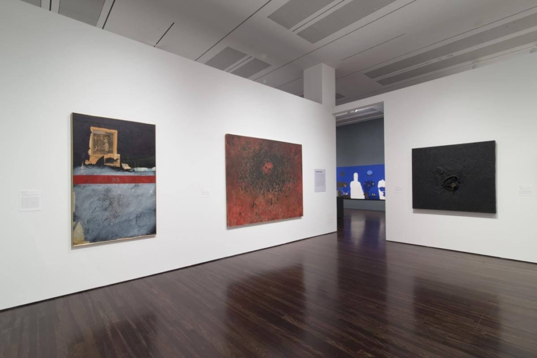 An art gallery with three abstract paintings displayed on white walls. The floor is dark wood, and the ceiling has recessed lighting. The left painting features a mix of dark and light colors with a red horizontal stripe. The middle painting is predominantly red with a dark, textured center. The right painting is mostly black with rough texture. A doorway in the background leads to another room with blue walls and white silhouettes.