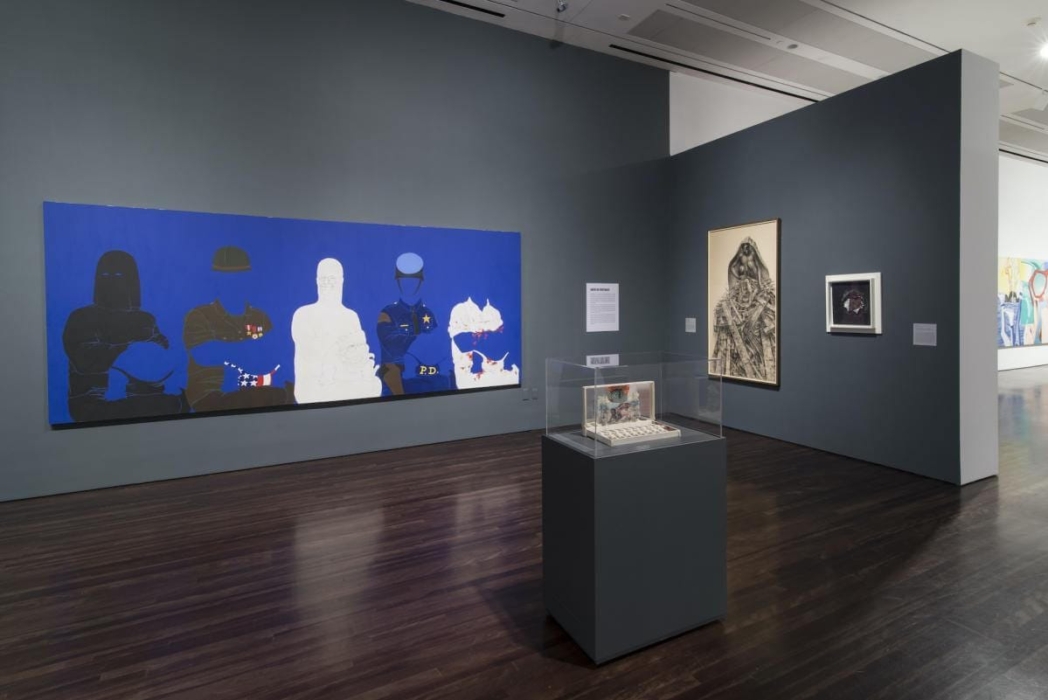 An art gallery featuring a large painting with five figures in various uniforms on a blue background, a glass case with a detailed artwork in the center, and two framed artworks on the right wall. One is a drawing of a figure wrapped in cloth or bandages, and the other is a smaller piece with a central figure on a dark background. A text panel on the right wall provides information about the art.