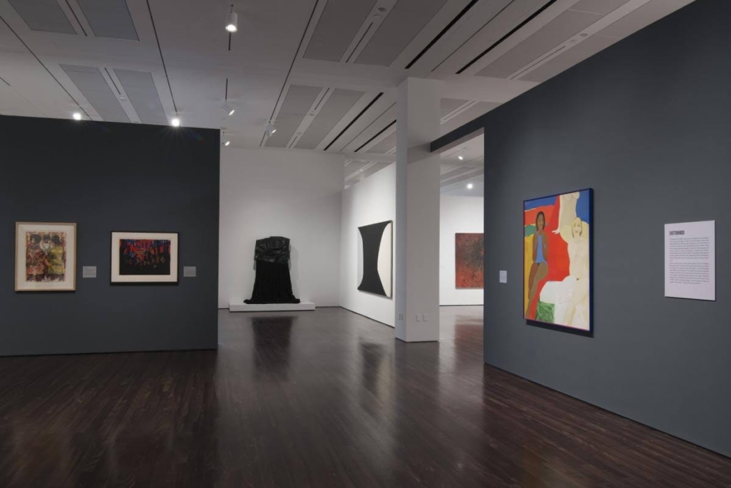 An art gallery with various artworks displayed on the walls. The gallery has dark wooden floors and a modern, minimalist design with white and dark grey walls. On the left wall, there are two framed artworks—one abstract painting with a mix of colors and a darker, more abstract piece. In the center of the room, there's a black sculpture on a white pedestal. On the right wall, there's a large, colorful painting depicting two figures, one with darker skin and the other with lighter skin, next to a text panel.