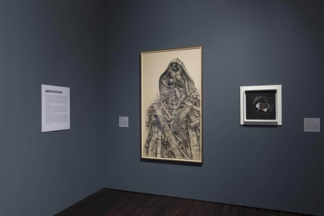 The image shows an art exhibition with two framed artworks displayed on a dark gray wall. The larger artwork on the left features a detailed drawing of a figure with a hood, surrounded by a chaotic arrangement of wooden planks or debris. The smaller artwork on the right appears to be a mixed media piece with a dark background and a central, textured element that looks like a torn or broken surface. To the left of the larger artwork, there is a text panel titled 'AMERICAN NIGHTMARE.' The text on this panel is not fully legible in the image, but it appears to provide context or information about the exhibition or the specific artwork. There are also small labels next to each artwork, likely providing the title, artist's name, and other relevant details about the pieces.